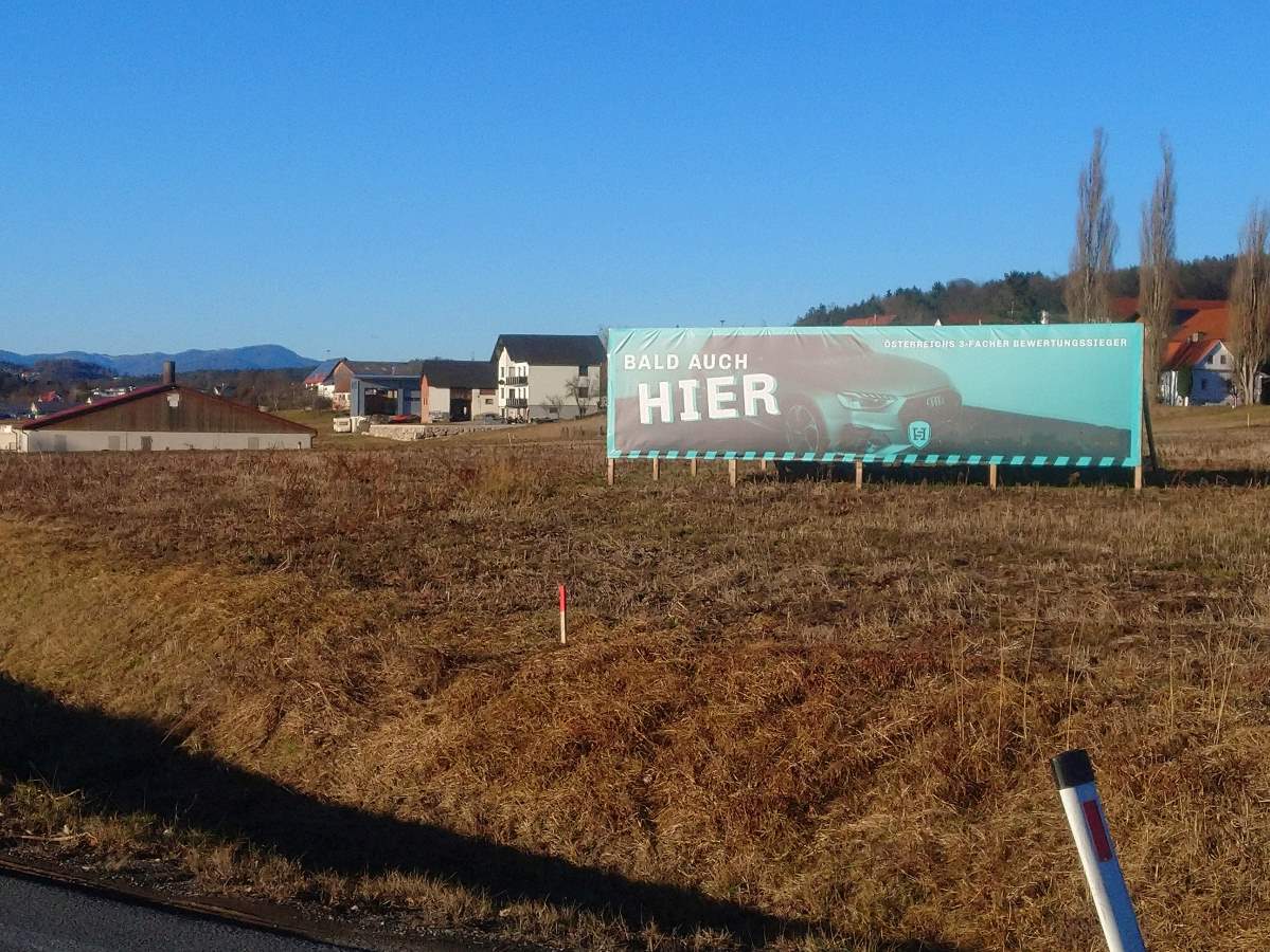 Zu sehen ist: ein Acker mit einem Plakat darauf. Text: Bald auch hier. Bild: ein Auto.