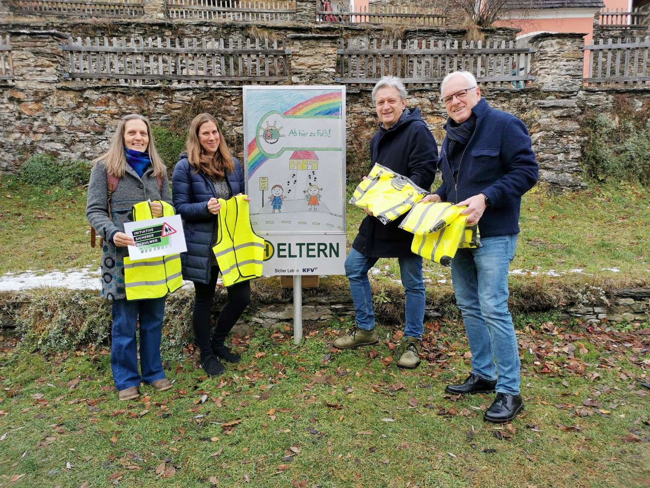 Zu sehen ist: Vier Personen stehen mit Warnwesten vor einer Schule