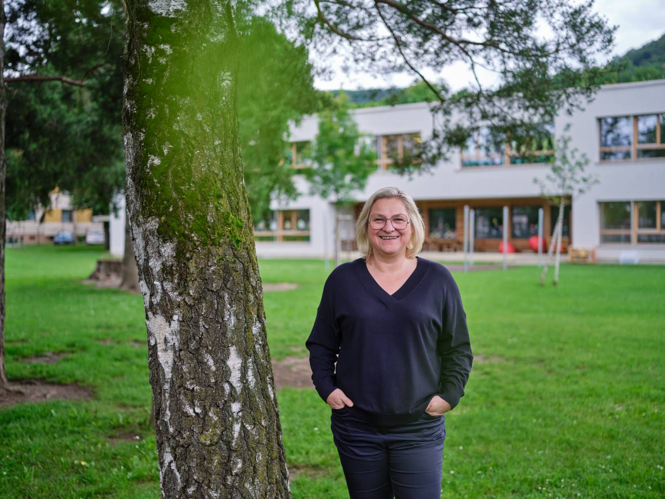 Zu sehen ist: eine Person steht vor einer Volksschule neben einem Baum